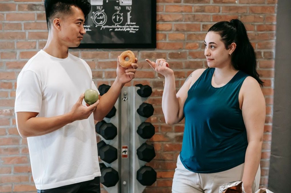 Side view of Asian personal instructor with apple and doughnut offering healthy food to plus size woman while standing in gym